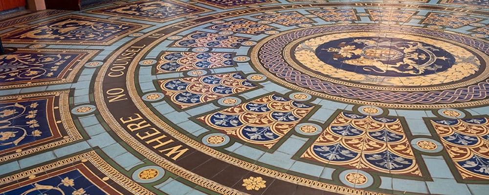 An ornate round mosaic floor with blue, gold and red tiles, and a crest in the middle. 